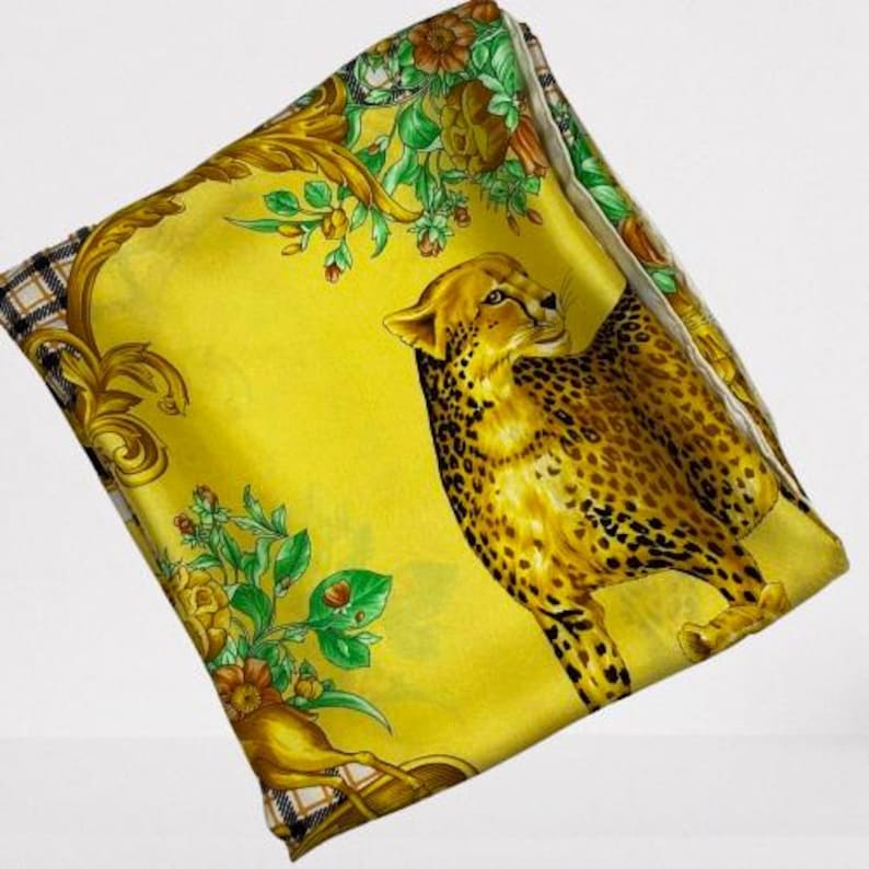 May include: Yellow silk scarf with a cheetah design. The scarf features a detailed illustration of a cheetah with brown spots, set against a yellow background. Floral and gold baroque accents frame the cheetah.