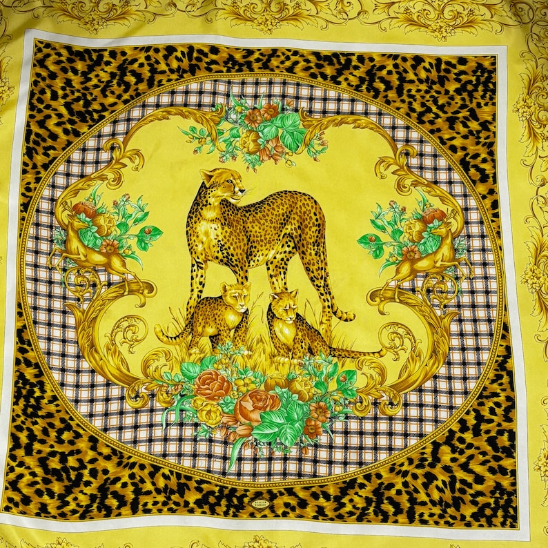 May include: A square yellow scarf with a cheetah family design. The center features a cheetah with two cubs, surrounded by floral and gold scrollwork. The border has a black and white checkered pattern and a leopard print.