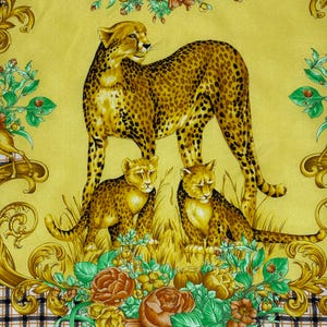 May include: A yellow scarf featuring a cheetah and two cubs in a grassy field, surrounded by floral and ornate gold accents. The cheetah has a spotted coat, and the cubs are smaller versions of the adult. The background is a solid yellow.