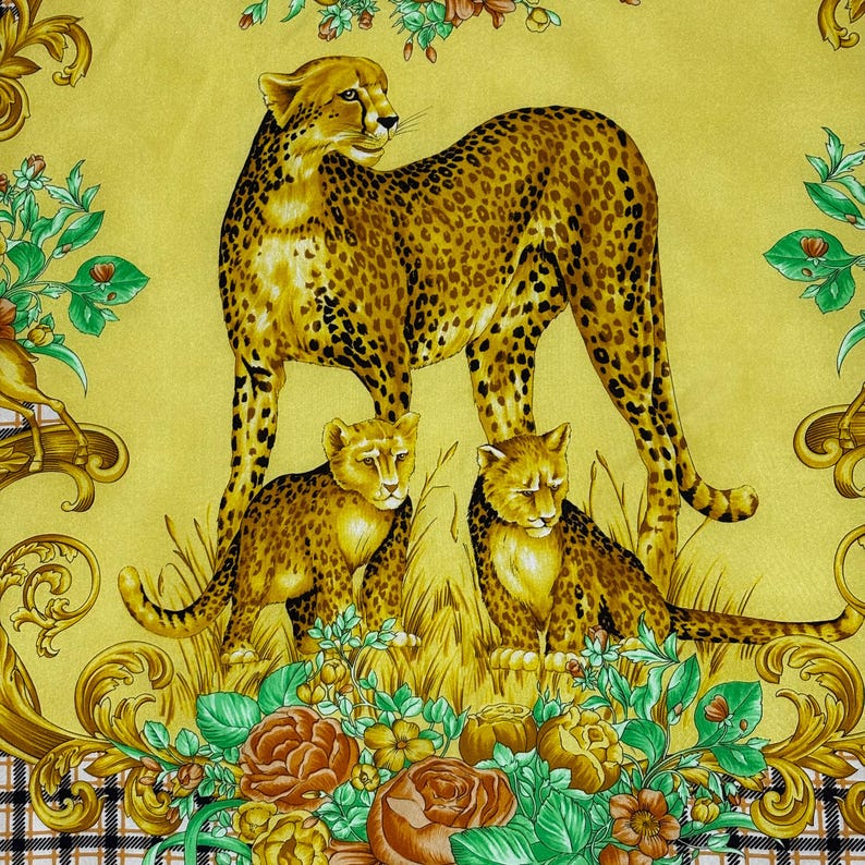 May include: A yellow scarf featuring a cheetah and two cubs in a grassy field, surrounded by floral and ornate gold accents. The cheetah has a spotted coat, and the cubs are smaller versions of the adult. The background is a solid yellow.