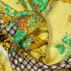 May include: A yellow silk scarf with a floral and animal print design. The scarf features roses, green leaves, and a leopard print pattern. The Atelier Versace logo is visible on the scarf.