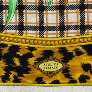 May include: Close-up of a silk scarf with a plaid pattern in black, white, and tan. The bottom section features a leopard print in black and gold, with the words "ATELIER VERSACE" in a diamond-shaped emblem.