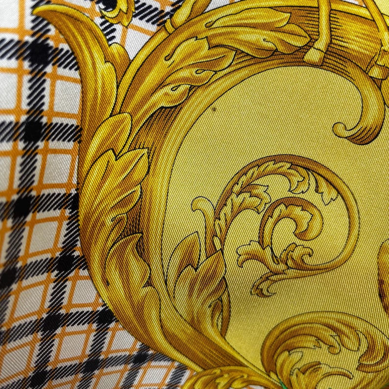 May include: Close-up of a silk scarf with a gold baroque design on a yellow background. The scarf features a plaid pattern in black, white, and orange. The intricate gold detailing includes ornate leaves and scrollwork.