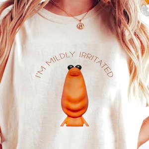 May include: Cream-colored t-shirt featuring a cartoon character with a long, orange body, black eyes, and small arms and legs. The text "I'M MILDLY IRRITATED" is arched above the character. The shirt is a casual, unisex style.