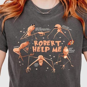 May include: Dark gray t-shirt featuring a cartoon graphic with the text "ROBERT HELP ME". The graphic includes several insect-like characters with speech bubbles. The shirt has a crew neck and short sleeves.