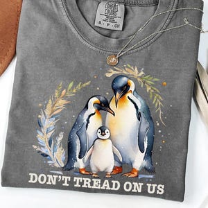 Könnte beinhalten: Ein graues Comfort Colors T-Shirt mit einem Pinguinfamilien-Motiv. Das Design umfasst zwei erwachsene Pinguine und ein Küken, mit dem Text "DON'T TREAD ON US" unten. Eine goldene Halskette ist über dem Shirt drapiert.