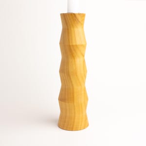 May include: A light-colored wooden candlestick with a geometric design. The candlestick has a cylindrical shape with a series of angled cuts creating a zig-zag pattern. A white candle is inserted at the top. The wood grain is visible.