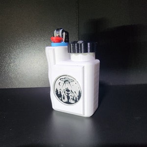 May include: White plastic container with a black lid and a red and blue lighter attached. The container has a sticker with a black and white illustration of an alien standing in a forest with a UFO above.