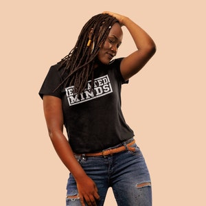Puede incluir: Una mujer con largas rastas marrones lleva una camiseta negra con el texto "Elevated Minds" impreso en ella. También lleva pantalones vaqueros azules con detalles desgastados y un cinturón marrón.
