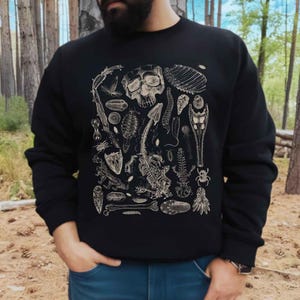 May include: Black sweatshirt featuring a detailed beige illustration of various anatomical elements, including a skull, bones, and insect parts. The design is centered on the front of the long-sleeved top. The person is wearing blue jeans.