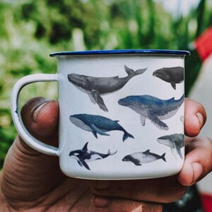 Peut inclure: Une tasse en &eacute;mail blanc avec un bord bleu, orn&eacute;e d'un motif de diff&eacute;rentes baleines dans des tons de gris et de noir. La tasse est tenue par une main devant une tente rouge et blanche.