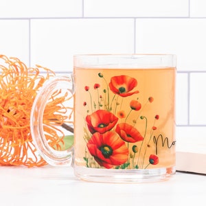 May include: A clear glass mug filled with a light orange liquid, decorated with red poppy flowers and the word "Mom". An orange flower and a small notebook are next to the mug against a white tile background.