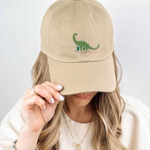Może przedstawiać: Beżowa czapka z daszkiem z wyhaftowaną grafiką zielonego dinozaura na środku z przodu. Czapka ma zakrzywiony daszek i usztywnianą koronę, prezentując prosty design z małą, szczegółową ilustracją przedstawiającą dinozaura z długą szyją.