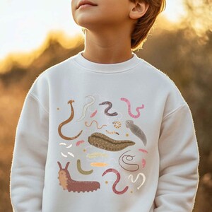 May include: White sweatshirt with a colourful illustration of worms and worm-like creatures. The design includes brown, pink, and tan worms, along with other shapes. The sweatshirt is worn by a person.