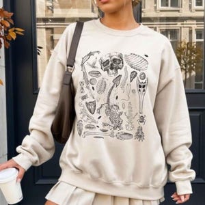Puede incluir: Sudadera beige con una ilustración anatómica en blanco y negro que presenta un cráneo, huesos y varios elementos biológicos. La sudadera se combina con una falda plisada a juego y botas blancas. Un bolso de hombro marrón y una taza de café blanca completan el conjunto.