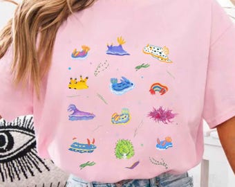 May include: A light pink t-shirt featuring colorful illustrations of sea slugs. The sea slugs are in various shapes and colors, including blue, yellow, purple, and green. The shirt is made of a soft material and has short sleeves.