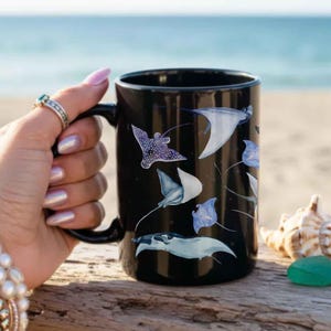 May include: A black ceramic mug featuring a repeating pattern of various stingrays in shades of white, blue, and grey. The mug has a cylindrical body and a curved handle on the side, showcasing detailed illustrations of marine life across its surface.