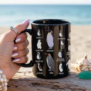 May include: Black ceramic mug featuring illustrations of various penguin species, including Humboldt, Emperor, and King penguins. The mug has a black handle and is held against a blurred beach background.