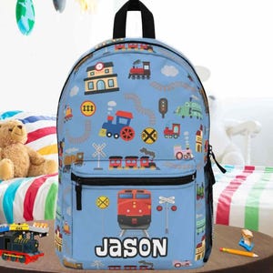 May include: A light blue backpack featuring a repeating pattern of colorful trains, railroad crossing signs, and buildings. The front pocket displays a large red train engine graphic above the name JASON printed in bold white capital letters.