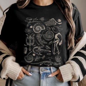 May include: A black short-sleeved t-shirt featuring a white graphic print of various prehistoric fossils, including ammonites, skeletal remains, and leaf imprints arranged in a dense, detailed collection across the center of the chest.