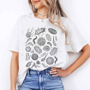 Puede incluir: Camiseta blanca con una ilustración en blanco y negro de varios microorganismos. El diseño incluye diferentes formas y tamaños de bacterias y virus. La camiseta se combina con unos vaqueros azules.