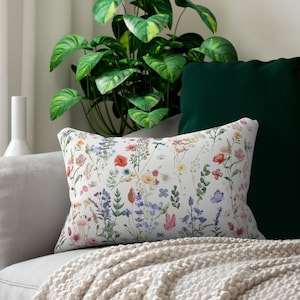May include: A rectangular throw pillow featuring a white background with a colorful botanical print of various wildflowers, grasses, and small butterflies in shades of red, blue, yellow, pink, and green, arranged in a scattered, natural pattern.