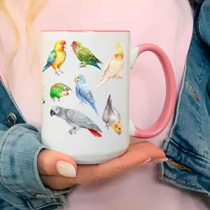 May include: A white ceramic mug with a pink handle and interior, featuring a colorful illustration of various parrot species including a sun conure, a green parrot, a cockatiel, a blue budgie, a grey parrot with a red tail, and a small green bird.