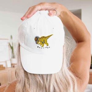 Pode incluir: Um boné de beisebol branco com um gráfico bordado de um dinossauro paquicefalossauro amarelo e castanho. O dinossauro é representado de perfil, virado para a esquerda, com pequenas pedras cinzentas espalhadas sob os pés na frente do boné.