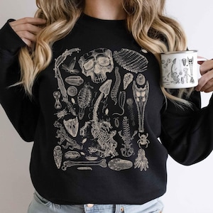 Op de afbeelding: Een zwart sweatshirt met lange mouwen en een gedetailleerde beige grafische print van diverse biologische specimens, waaronder een schedel, skeletstructuren, insecten en fossielen in een rasterpatroon op de voorzijde.