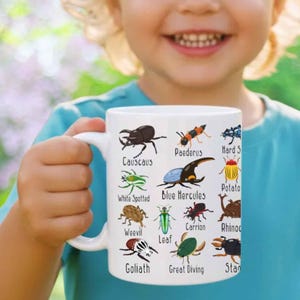 Peut inclure: Mug en céramique blanche orné d'illustrations colorées d'insectes, notamment des coléoptères et des charançons, avec leurs noms imprimés en dessous. Le mug est tenu par une personne portant un t-shirt bleu. Le mug est de taille standard.