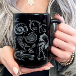 Può includere: Tazza in ceramica nera con illustrazioni bianche di vari fossili, tra cui ammoniti e resti scheletrici. La tazza ha un manico nero. Un regalo unico per gli appassionati di scienza. La tazza è tenuta da una persona.