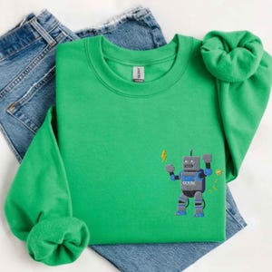 Puede incluir: Una sudadera verde brillante con cuello redondo y mangas largas. Presenta un diseño de robot bordado en gris, azul y negro, con rayos amarillos. La sudadera se combina con unos vaqueros azules.