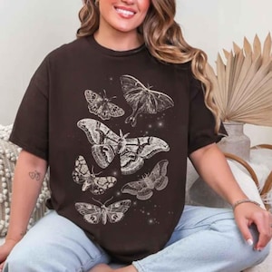 May include: A dark brown short-sleeve t-shirt featuring a white graphic print of five different moths arranged in a cluster. The design includes small white star-like dots scattered among the moths, creating a celestial effect on the front of the shirt.