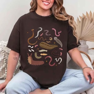 Peut inclure: Un t-shirt marron foncé à manches courtes avec une impression graphique représentant divers vers et créatures ressemblant à des limaces dans des tons rose, jaune, blanc et marron. Le motif est centré sur la poitrine et montre plusieurs espèces.