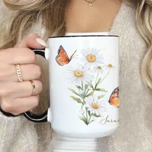 May include: White ceramic mug with a black handle and rim, decorated with watercolor daisies and orange butterflies. The mug has the name "Sara" written in script. A decorative floral mug.