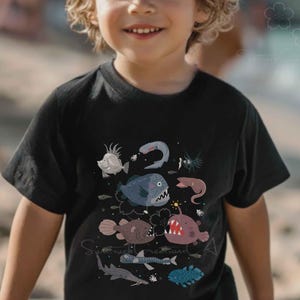 May include: A black short-sleeved t-shirt featuring a graphic print of various deep-sea creatures, including an anglerfish with a glowing lure, a shark, and other marine life in shades of blue, grey, and red, arranged in a scattered pattern.