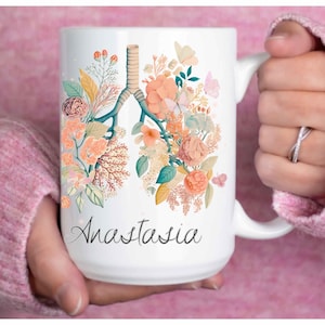 Puede incluir: Una taza de cerámica blanca con una ilustración colorida de pulmones humanos compuesta por varias flores y follaje en tonos naranja, melocotón y verde. El nombre Anastasia está impreso en una fuente cursiva negra bajo el diseño floral.