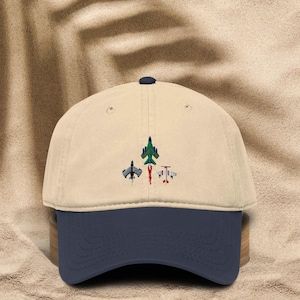 May include: A beige baseball cap with a navy blue brim. The cap features an embroidered design of three stylized airplanes in green, black, and red. The cap is made of a soft, durable fabric.