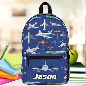 May include: A blue backpack featuring a repeating pattern of various aeroplanes in white, red, and orange. The front pocket displays a large white aeroplane graphic above the name Jason printed in bold white letters. It has a black top handle and zippers.