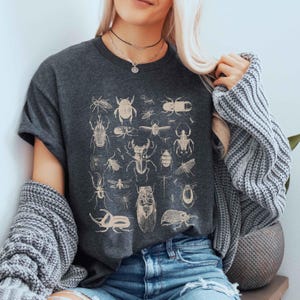 May include: Dark grey t-shirt with a beige insect illustration. The graphic features various beetles, bugs, and insects. The shirt has short sleeves and a crew neck. The overall theme is nature and entomology.