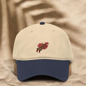 May include: A beige baseball cap with a dark blue brim and a small embroidered graphic of a brown and red trilobite creature on the front panel. The cap features a dark blue button at the top centre and structured stitching across the crown.