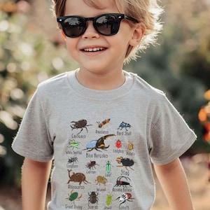 Peut inclure: Un t-shirt gris clair à manches courtes avec une impression graphique colorée représentant divers insectes et coléoptères. Chaque insecte est étiqueté avec son nom, notamment Blue Hercules, Rhinoceros, Stag et Goliath, disposés en grille.