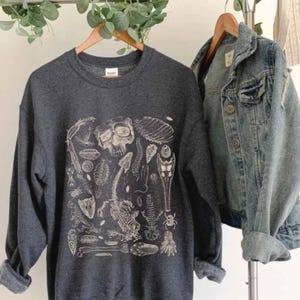 Op de afbeelding: Een donkergrijze sweater met ronde hals en een witte grafische print van diverse gefossiliseerde skeletresten, waaronder een schedel, ruggengraat en insect, gepresenteerd naast een blauw spijkerjack op een kledingrek.