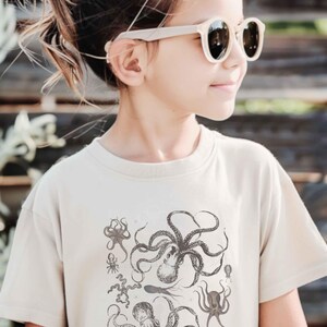 May include: A beige short-sleeved crew neck t-shirt featuring a detailed black ink-style graphic print of various octopus species and marine life arranged in a cluster on the front center of the garment.