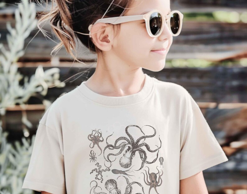May include: A beige short-sleeved crew neck t-shirt featuring a detailed black ink-style graphic print of various octopus species and marine life arranged in a cluster on the front center of the garment.