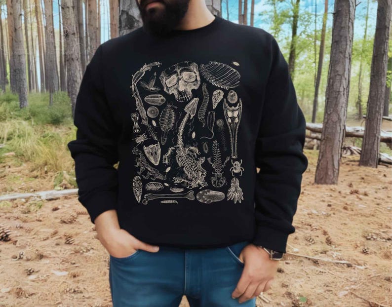 May include: Black sweatshirt featuring a detailed beige illustration of various anatomical elements, including a skull, bones, and insect parts. The design is centered on the front of the long-sleeved top. The person is wearing blue jeans.