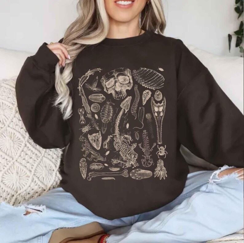 Op de afbeelding: Een donkerbruin sweatshirt met ronde hals en een beige grafische print van diverse anatomische skeletstructuren, waaronder een menselijke schedel, wervelkolommen, ribbenkasten en diverse biologische specimens.