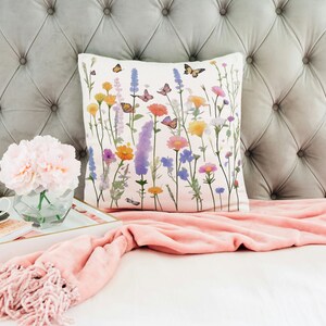 May include: A white throw pillow with a floral print featuring colorful flowers and butterflies. The pillow is on a gray tufted headboard with a pink throw blanket.