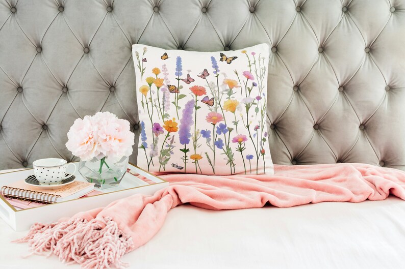 May include: A white throw pillow with a floral print featuring colorful flowers and butterflies. The pillow is on a gray tufted headboard with a pink throw blanket.