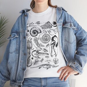 May include: A white t-shirt featuring a black ink graphic print of various fossils, including ammonites, a turtle shell, a skull, and prehistoric skeletal remains, worn under a light blue denim jacket with metal buttons and chest pockets.